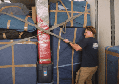 A man is securing a padded furniture load with logistic straps inside a moving truck